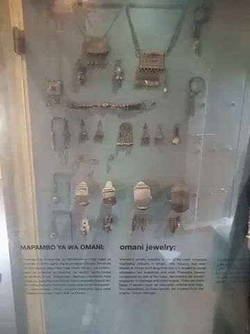 The video shows a museum display case featuring Omani jewelry and traditional Omani women's clothing. The jewelry exhibit displays various silver necklaces, earrings, and pendants. Next to the jewelry display are several mannequins dressed in Omani women's clothing. The clothing varies in color and design, including dresses in pink, blue, black and gold. The mannequins also wear headscarves.