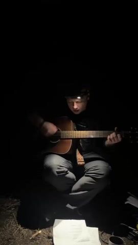 A medium shot shows a young man sitting cross-legged in the dark, illuminated by the headlamp he is wearing. He is strumming a brown acoustic guitar. Sheets of music are at his feet, as is a mug. He looks down at the guitar as he plays.