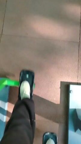 The video shows a first-person perspective of someone using a piece of gym equipment. The person is wearing dark pants and light-colored shoes, which are placed on black foot pedals with green accents. The foot pedals are moving up and down, indicating the person is stepping or pedaling. The gym floor is a reddish-brown color, possibly a rubberized mat. To the left and right of the foot pedals, there are parts of the gym equipment that appear to be made of grey metal and possibly blue plastic. The lighting suggests an indoor environment, with some shadows cast on the floor. The camera angle is looking down at the person's feet and the equipment.