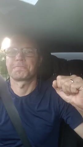 The video shows a bald man with glasses wearing a dark blue t-shirt and a seatbelt. He is in the passenger seat of a car. The sun is shining brightly through the windshield, causing lens flare. The man holds a white object in his left hand and makes a fist with his right hand. He appears to be singing or talking along to music. The background is blurred due to the motion of the car.