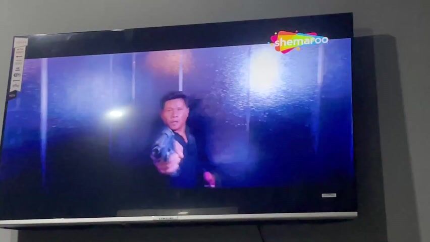A television screen is mounted on a gray wall, displaying a scene from a movie or show. The black Samsung TV has a silver trim along its bottom edge.

On the TV screen, a man in a dark shirt is visible, holding a handgun and pointing it forward. The scene has dramatic lighting with blue and purple hues, possibly set in an elevator or a modern interior with metallic or glass surfaces. Another person's hands are briefly visible in the foreground of the TV screen, appearing to be in an action sequence or struggle.

In the top right corner of the TV screen, the colorful "shemaroo" logo is prominently displayed, indicating the content provider. On the left side of the TV, several small icons and text like "4K", "Wi-Fi", and "NEW" are visible, likely part of the TV's display features or branding. The image is focused on the television screen, capturing the ongoing action sequence. The video was recorded in Rasoolpur, Pakistan.