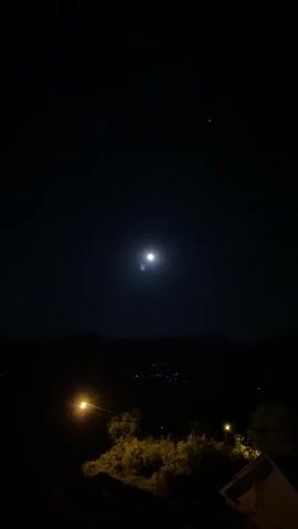 The video shows a nighttime view of the sky with a bright, full moon. There are some trees and buildings silhouetted in the foreground. In the far distance, there are small lights that could be from a town or city. A sound of someone singing is heard in the background.