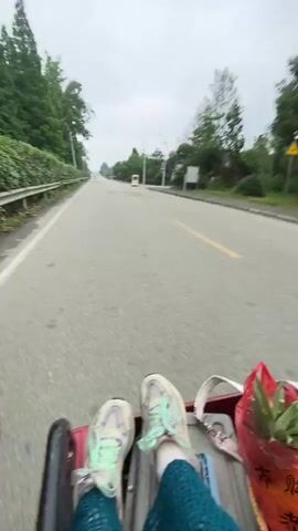 This video appears to capture a ride in a three-wheeled vehicle, possibly an auto-rickshaw, on a cloudy day in Chongzhou City, China. The scene is set outdoors, with greenery and a road visible in the background.

The primary subjects are three young women. The person closest to the camera, with curly blonde hair and wearing a green hooded jacket and a pink furry purse, is likely the one filming or at least capturing the selfie-style footage. She looks directly at the camera with a neutral or slightly amused expression. To her left, a woman with dark hair, glasses, and wearing a purple top is looking down at her phone. Behind the blonde-haired woman, another woman with dark hair, glasses, and wearing a black top is seen driving the vehicle. Her gaze is directed forward, focusing on the road.

The activity taking place is a journey on this motorized three-wheeler. The vehicle has a canopy, suggesting it might be used for transportation in varying weather conditions. The wind is noticeable, as it blows through the blonde woman's hair.

Notable details include the overcast sky, indicating possibly a mild or humid day, and the presence of trees and a road, typical of a semi-urban or rural setting. The women appear relaxed and engaged in their own activities while traveling. There is some writing visible on the side of a white truck in the background of the first frame, but it is too blurry to be legible. In the second frame, there are some Chinese characters on the canopy of the vehicle, but they are also difficult to read clearly.
