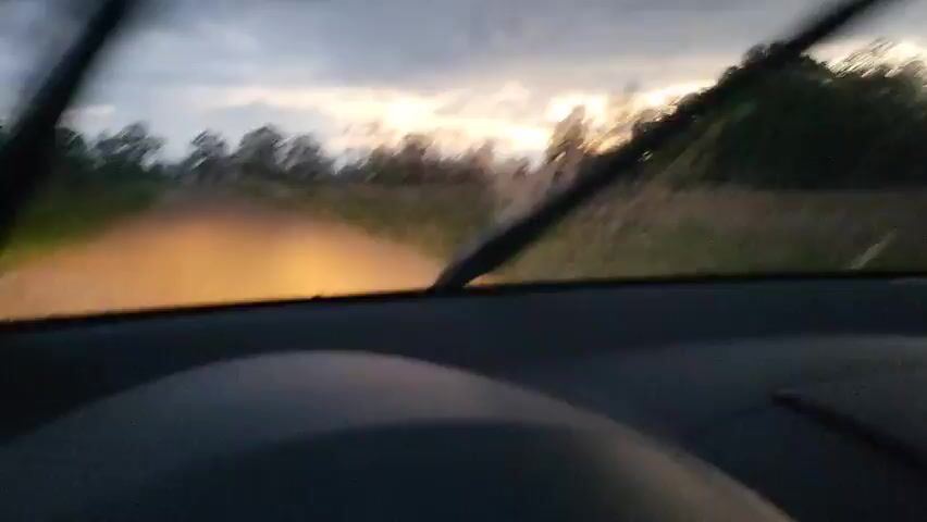 The video shows the view from inside a car driving down a rural road during what appears to be a storm. The windshield wipers are active, clearing rain from the glass. The sky is overcast with patches of sunlight breaking through. Trees and fields are visible on either side of the road. The headlights of the car are on. The interior of the car is dimly lit, with the dashboard and steering wheel visible in the foreground.