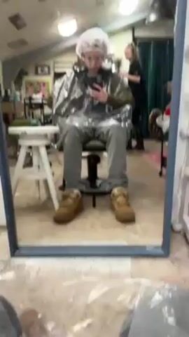 A young person in a hair salon is seated in a chair, wearing a protective plastic cape and a hair net, and taking a selfie in a mirror. The person is wearing gray jeans and tan work boots. In the background, a woman is working on someone's hair. The salon appears well-lit with overhead lights, and there are various salon supplies and decorations around the room, including a mannequin head with a dark wig, a white ladder shelf full of products, and artwork on the walls. The person taking the selfie is holding a dark-colored smartphone. The camera pans slightly, offering a wider view of the salon setting.