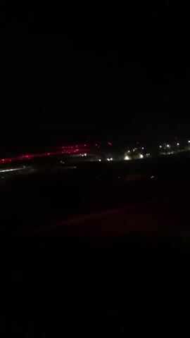 The video shows an airplane taxiing on a runway at night. The runway is lit with red and white lights. Other lights are visible in the distance, likely from the airport terminal or surrounding buildings. The camera appears to be positioned inside the airplane, looking out the window.