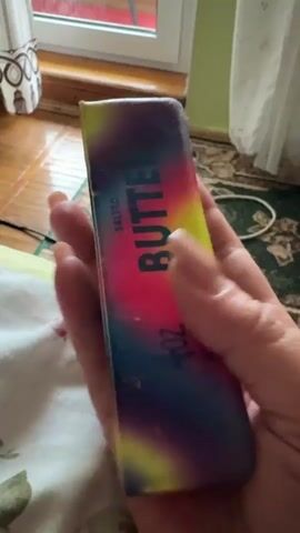 A hand holds a squishy stress ball with a rainbow color pattern and the word "BUTTER" printed on it. The hand squeezes the stress ball, distorting its shape. The background is out of focus but appears to be a room with a window and a carpet.