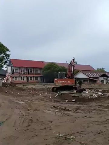 The video shows a construction site with an excavator and a dump truck. The ground is muddy and appears to be a cleared area. In the background, there are buildings that look like they could be homes or a school, and a dome that resembles a mosque. The sky is overcast. There is a person sitting on the ground near the excavator. A motorcycle is parked to the left of the dump truck. The dump truck is in the process of dumping its load of dirt. The scene suggests ongoing demolition or land clearing work.