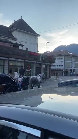 A horse-drawn carriage is on a street in front of buildings. A driver is on the carriage, and there are passengers. The sun is setting, and the sky is light blue with some clouds. The buildings appear to be old and made of brick. There are cars on the street, and the foreground is blurry, suggesting the video was taken from a moving vehicle.