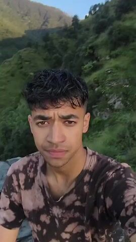 The video is a first-person perspective selfie video taken outdoors. The subject is a young man with curly dark hair, wearing a tie-dye t-shirt and dark shorts. He is sitting on rocks with a lush green mountainous landscape and a town nestled in the valley behind him. The sky is partly cloudy and appears to be during the day. He shifts his pose slightly, turning his head to look at the camera. The video is brief and appears to be a quick recording of the scenic view.