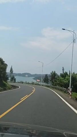 The video shows a car driving on a winding road next to a lake. The road is paved and has yellow lines marking the center. There is a guardrail along the side of the road. On the other side of the guardrail is a view of a lake with green hills and vegetation. There is also a man walking on the side of the road. The sky is blue with some clouds.