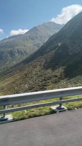 A scenic video captures a mountain landscape from a moving vehicle. The camera pans across a vast, rugged mountain slope covered in rocks and sparse vegetation, with a clear blue sky and scattered clouds overhead. In the distance, a winding road can be seen snaking through the valley, with a few small buildings visible along its route. The video appears to be filmed on a sunny day, offering a glimpse of a picturesque mountain pass.