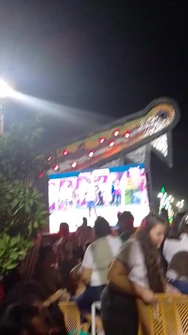 The video shows a crowd of people at night, seemingly at an outdoor event or concert. There's a large screen displaying an image or video, along with a lit-up structure in the background. The crowd is dense and individuals are holding up phones, possibly taking photos or videos of the event. The lighting is dim, relying on the screen and structure for illumination. The camera is moving, giving a sense of the viewer navigating through the crowd.