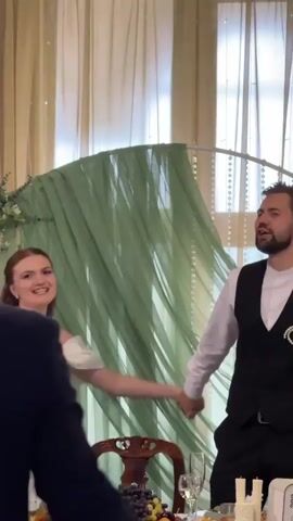 A wedding reception is in progress. A bride in a white off-the-shoulder wedding dress and a groom in a black vest and white shirt are holding hands and standing at a table laden with food, including fruit, and bottles of wine. The bride smiles and looks towards the groom, who is looking at her and speaking. Behind them, a draped green fabric is suspended from an archway. The room is lit and appears to be indoors. The video is a close-up, handheld shot.