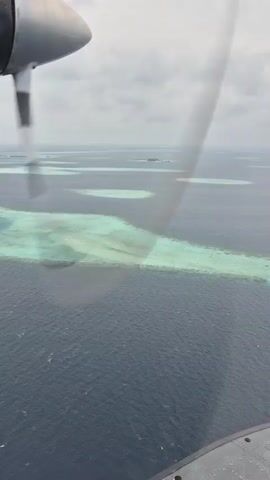 The video shows an aerial view of a seascape, likely in the Maldives, as seen from a small airplane. The predominant colors are the deep blue of the ocean and patches of turquoise indicating shallow waters or coral reefs. Several small, sandy islands are scattered across the horizon under a cloudy sky. A portion of the airplane's wing and engine are visible in the foreground, displaying branding for "NH Collection Hotels & Resorts" and another logo that partially reads "Wani." The propeller is rotating, contributing to the overall dynamic perspective of the footage.