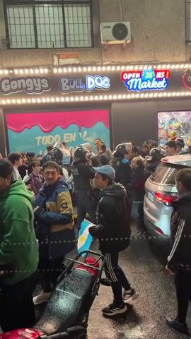 The video shows a crowded street scene at night. The crowd appears to be gathered in front of a store with illuminated signage that reads "Gongy's," "Bull Dog," and "Open 24 hs Market." The store's facade is decorated with colorful lights. In the foreground, a silver Jeep is parked, and several people are standing around it and walking past. Some individuals are wearing what appear to be football jerseys, and one person is holding an Argentinian flag. The ground appears wet, suggesting it may have rained. The overall atmosphere seems to be one of excitement and gathering, possibly related to an event or celebration.