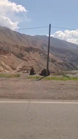 The video pans from left to right across a landscape featuring a river snaking through a valley with mountains on either side. The sky is blue with scattered white clouds. The mountains appear dry and rocky. The river is wide and shallow. The camera seems to be positioned on a road running parallel to the valley. A power pole is visible at the beginning of the video. A voice can be heard speaking in a foreign language, possibly exclaiming about the beauty of the scenery.