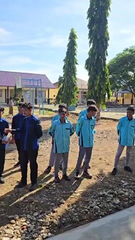 A group of young people, most likely students, stand outside near buildings. The people are dressed in matching uniforms. Some wear light blue shirts and grey pants, while others wear dark blue shirts and grey or black pants. Some of the individuals are wearing headscarves. In the background, there are school-like buildings with red roofs and yellow walls. There are also tall, thin trees, and a flag pole with a flag. The ground is rough with stones and dirt. The sky is blue with some clouds. The scene appears to be during the daytime, possibly a school assembly or gathering.