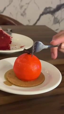 A fork is used to cut into a red, spherical dessert that is presented on a white plate with a light brown circular base. The dessert is cut in half, revealing a white interior. To the left of the dessert, on another white plate, is a slice of red velvet cake. The background shows a wooden table and a marble-patterned wall.