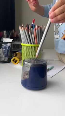A close-up shot shows a child's hands sharpening a pencil in a blue manual pencil sharpener. The child is wearing a light blue long-sleeved shirt with a cartoon character on it. In the background, a green metal cup holds a collection of pencils, and other art supplies are visible on a white desk. The video captures the action of sharpening the pencil in a steady, focused manner.
