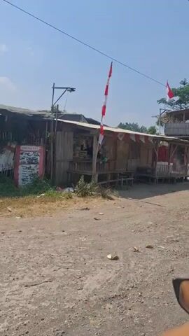 The video opens with a view of a roadside structure with a corrugated metal roof, wooden walls, and a raised wooden platform. Red and white flags, resembling the Indonesian flag, are visible on the structure and on a taller wooden tower behind it. Logs are stacked beside the tower. The ground in front of the structure is dirt. The camera then pans to the right, showing a road with trees and vegetation alongside it. A motorcycle is seen riding on the road in the distance. The sky is clear and blue. The scene appears to be in a rural or semi-rural area.