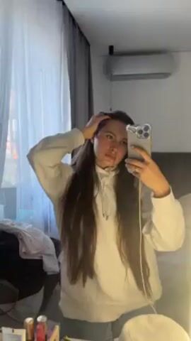 A woman with long brown hair and wearing a white hoodie stands in front of a mirror, holding a smartphone to take a selfie. She is in what appears to be a hotel room or bedroom. There is a bed behind her and a window with curtains to the left. An air conditioning unit is mounted on the wall. Personal items are visible on a surface near the mirror. The woman smiles slightly as she looks at the camera. She runs her hand through her hair.