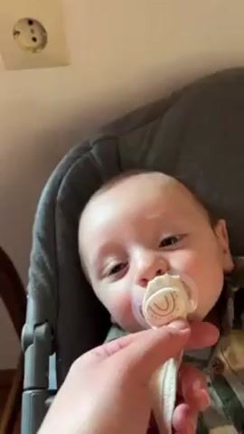 Here is a description of the video:
The video shows a baby sitting in a gray high chair, wearing a plaid shirt. An adult's hand holds a pacifier to the baby's mouth, and the baby takes it. The baby has blue eyes and fair skin. The background includes a white wall with an electrical outlet and part of a wooden table. The lighting is bright.