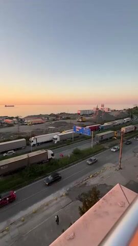 The video captures a wide shot of a coastal area during sunset. The sky is a gradient of orange and pink, transitioning to clear blue. The sea stretches out to the horizon, where a large ship is visible. Along the coastline, there are several industrial-looking structures and stacks of what appear to be construction materials or gravel. A busy road runs through the scene, with multiple trucks lined up, suggesting a traffic jam or a waiting area for transport. Several cars are also on the road, moving in both directions. In the foreground, a portion of a building's exterior, possibly a balcony or railing, is visible. A single person is walking on the sidewalk in the lower left corner of the frame. The overall impression is of a functional, possibly port or industrial, area at the end of the day.