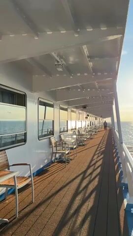 The video captures a view from the deck of a boat or ship. The deck is made of dark wood and lined with tables and chairs, suggesting an outdoor seating area. The side of the boat, painted white with a blue stripe, features several windows. The sun is low in the sky, casting a warm glow and creating long shadows on the deck, indicating either sunrise or sunset. The sea stretches out to the horizon, with gentle waves reflecting the sunlight. A single person is walking away from the camera down the deck, towards the sun. The overall atmosphere is peaceful and serene, with the sound of water and distant seagulls.