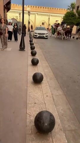 The video shows a street scene in what appears to be a city in Morocco. A modern white Mercedes car is parked on the left side of the street, with a row of black ball-shaped bollards separating the car from the sidewalk. On the right side of the street, a horse-drawn carriage is moving towards the viewer. The carriage is pulled by two horses, one white and one brown. There are people in the carriage, and the driver is waving. In the background, there are buildings with distinct Moroccan architectural features, including arched doorways and a tall minaret with patterned tiles. The sky is overcast, suggesting it is either late afternoon or early evening. The scene presents a contrast between modern transportation (the car) and traditional transportation (the horse-drawn carriage).