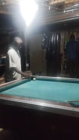 The video depicts a dimly lit, indoor space with a pool table as the central focus. A man in a light-colored shirt is positioned at the pool table, his hand near a ball. Other individuals, including a person in a red shirt, are visible in the background. The environment appears to be a casual setting, possibly a recreational area or bar. The lighting is uneven, with a bright overhead light dominating the scene. Overall, the video captures a moment of recreation or leisure activity involving a game of pool.