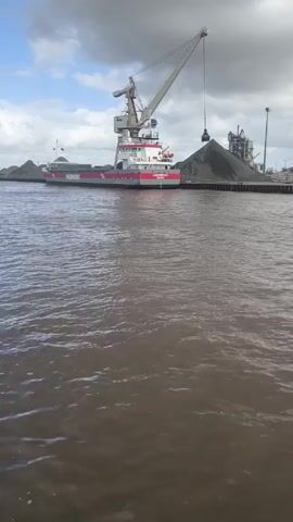 The video shows a large crane on a ship with the name "LAURINBORG" painted on its side, unloading a pile of dark material onto a dock. The ship has a red, white, and gray color scheme. Behind the pile of material on the dock, there is a structure with various tanks, pipes, and a smokestack. The sky is overcast, with patches of blue visible through the clouds. The water in the foreground is brown and choppy.