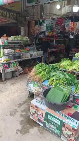 The video shows a market stall stocked with fresh produce. In the foreground, bunches of leafy green vegetables with roots are displayed. To the left, there are baskets of cabbage and other vegetables like carrots and green beans. In the middle, a man in a black shirt and blue jeans stands near the produce, appearing to be a vendor. Behind him, shelves are stocked with various packaged goods. Piles of coconuts are visible at the base of the stall. The environment appears to be an outdoor or semi-outdoor market with a concrete floor. The camera pans slightly, giving a view of the abundance of fresh food for sale.
