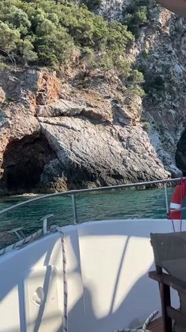 A boat is moving towards a cave in a rocky cliff face. The water is turquoise. The cliff is covered in greenery. The boat has a white hull and railing, a life preserver, and a chair.