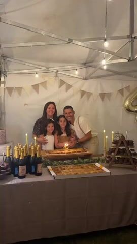 A family of four, two adults and two young girls, are gathered around a table for a celebration. The family is smiling and looking at the camera. There are two cakes on the table, both with lit candles. Several bottles of champagne are also on the table, along with a tiered display of cupcakes. The scene appears to be set up outdoors under a canopy, with decorative bunting strung across the back wall. The overall atmosphere is festive and celebratory.