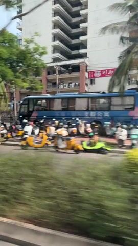 The video captures a view from a moving vehicle, likely a bus or car, passing by a street scene. Tall buildings with multiple floors and balconies are visible in the background. Trees and lush green foliage line the street. Parked motorbikes and scooters are visible along the side of the road. A blue bus is also seen at one point. The footage is slightly blurry due to the motion, suggesting a continuous travel or a rapid pan. The lighting suggests it is daytime.