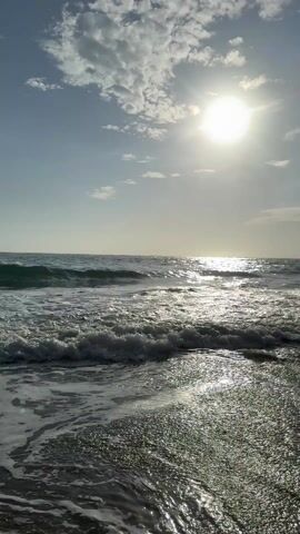 The video shows a sunny day at the beach. Waves are rolling in and breaking on the sand. The sun is bright and high in the sky, partially obscured by scattered clouds. The water is reflecting sunlight. The beach appears empty.