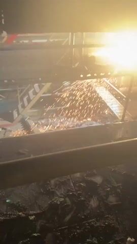 A worker on scaffolding is using a torch to cut metal, creating a shower of bright sparks that fall into a debris-filled pit below. The scene is dimly lit, with the bright sparks illuminating the area.
