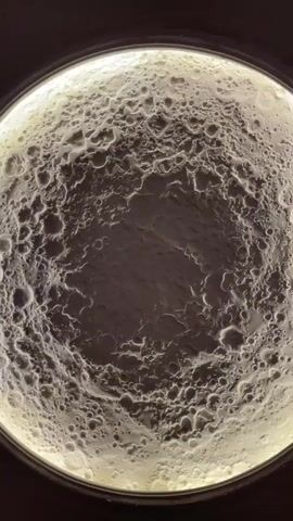 A close-up shot shows a circular object that resembles the surface of the moon. The surface is textured with craters and appears to be illuminated from behind, causing a bright glow around the edges. The center of the object is darker and has a rough texture. The camera angle is overhead, looking down at the object.
