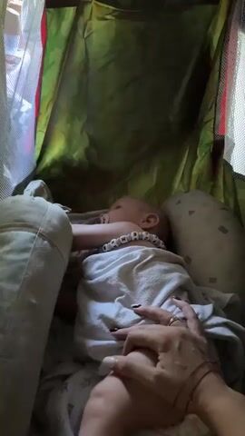 A baby is sleeping in a crib with green camouflage fabric. A person's hands are visible, holding the baby. Another person, a child, is looking into the crib from above. The scene appears to be indoors, with light filtering in from outside.