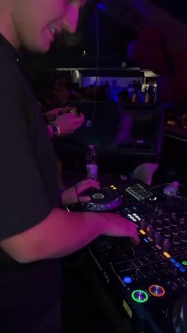 The video shows a DJ mixing music at a crowded event. The DJ is wearing a black shirt and is using a professional DJ mixer with turntables, knobs, and illuminated buttons. The crowd is diverse, mostly dressed in white shirts and suits, and is dancing with their hands in the air. There are red balloons decorating the ceiling. The lighting is dynamic, with purple and blue lights flashing across the room, adding to the energetic atmosphere.
