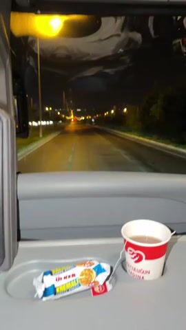 The video shows a close-up of a tray inside a bus or train at night. On the tray are a package of Ülker Kremalı biscuits and a cup of tea. The bus or train is moving, as indicated by the blur of lights and the road visible through the window. The exterior scene is dark, with streetlights and distant city lights. No people are visible in the frame. The overall mood is one of quiet travel during the night.