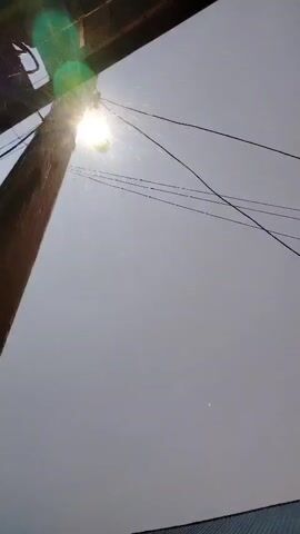A video captures a close-up view of a wooden pole with wires running across the frame. The sun shines brightly, causing lens flare and making it difficult to discern details. Specks of light, possibly dust or water droplets, are visible in the air. As the video progresses, the sky transitions from a bright, overcast gray to a gradient of pink and purple, suggesting either a sunrise or sunset. Below the wires and the pole, the roofline of a building is visible, with a dark, textured surface and what appear to be windows and a doorway. The camera angle is tilted upwards, looking towards the sky.