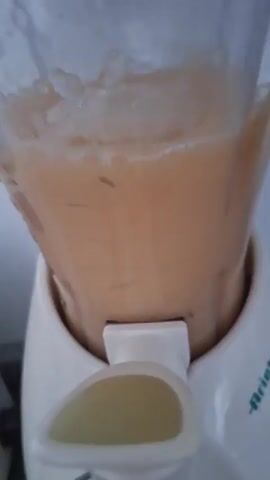 A stationary video shows a white Ariete smoothie maker containing a smoothie being briefly activated. The smoothie is light orange and contains dark specks. The machine is sitting on a grey countertop, with a white wall in the background. A clear glass is placed directly under the dispenser spout of the machine by a hand. The machine runs very briefly.