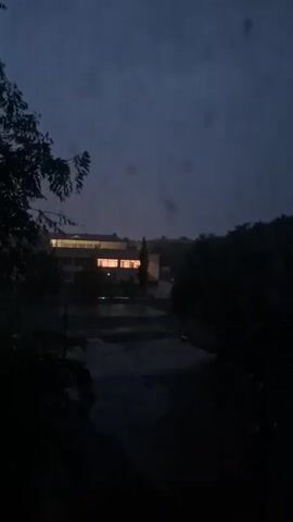 This is a short video showing a dark, overcast sky with some light reflecting off what appears to be rain or water. In the distance, a building with illuminated windows is visible. Trees and foliage are present in the foreground and to the sides, obscuring parts of the scene. The overall mood is somber and atmospheric, suggesting twilight or a storm.