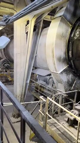 The video shows a large, cylindrical industrial machine, likely a kiln or dryer, in operation. The machine is covered in a white powdery substance, suggesting it processes materials like cement or lime. The camera is positioned on a metal walkway or platform overlooking the machinery. The environment appears industrial, with visible metal structures, railings, and lighting. The machine itself is massive and appears to be rotating, indicated by the slight blurring of its edges and the dust it seems to be shedding. There are no people visible in the video.