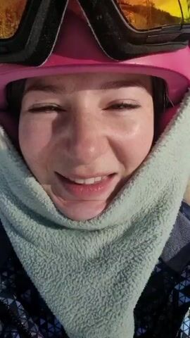A young woman is on a ski lift, smiling at the camera. She is wearing a pink helmet, ski goggles, and a gray neck warmer that frames her face. The sun is reflecting in her goggles. She is also wearing a dark jacket with a geometric pattern. The background shows a snowy mountain landscape with trees and a blue sky with some clouds. The ski lift cable and support structures are visible.