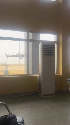 The video shows the interior of an airport terminal, focusing on a window view. Outside the window, an airfield with a helicopter taking off can be seen. The helicopter is gray, and appears to be a military model. Inside, there's a large, white air conditioning unit next to the window, and a row of airport seating is partially visible in the foreground. A "Dust Bin" sits below the window. The walls are painted a light yellow color. The weather outside appears overcast.