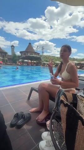 A woman in a white bikini sits on a lounge chair by a swimming pool. She holds a bottle of sunscreen and makes a peace sign with her fingers. In the background, people swim in the pool and lounge under umbrellas. A man with tattoos sits on a nearby lounge chair. The sky is blue with white clouds.