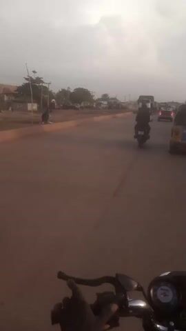 The video, shot from the perspective of a motorcyclist, depicts a busy road with vehicles and people moving in both directions. The road appears to be made of red dirt or soil. Motorcyclists, a truck, cars, and a yellow auto-rickshaw are visible. The truck, labeled "EUROTRAKKER," is white and carries a large covered load. The sky is overcast.  The overall environment suggests a developing or rural area.  There is a sign with "GAS" on it, possibly indicating a nearby gas station. The motorcyclist is stopped, apparently waiting for traffic to move.